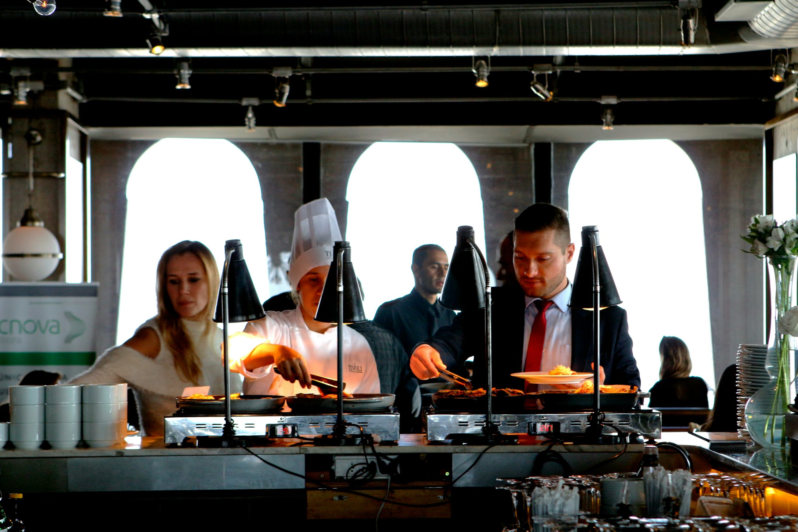 People serving food at a buffet in a restaurant. A chef in a white uniform and hat is preparing dishes, while a man in a suit and a woman in casual attire select their meals under warm lights.