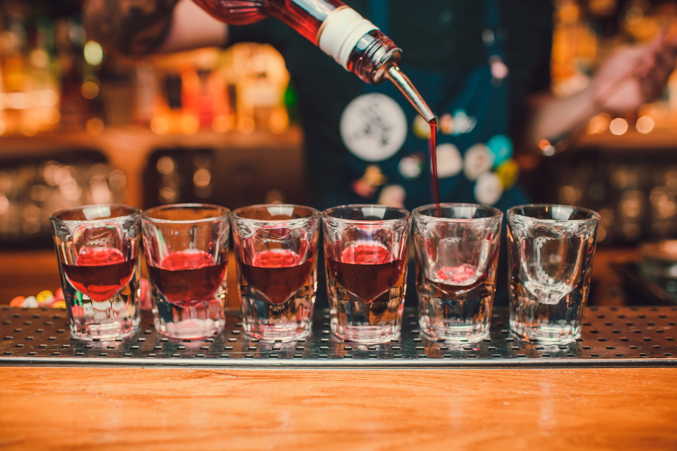 A bartender pouring red liquid into a series of shot glasses arranged on a bar counter, with a colorful and lively background of a bar environment. The bartender is wearing an apron adorned with pins and badges, adding a personalized touch to the scene. The focus is on the glasses and the act of pouring, capturing the essence of mixology and bar service.