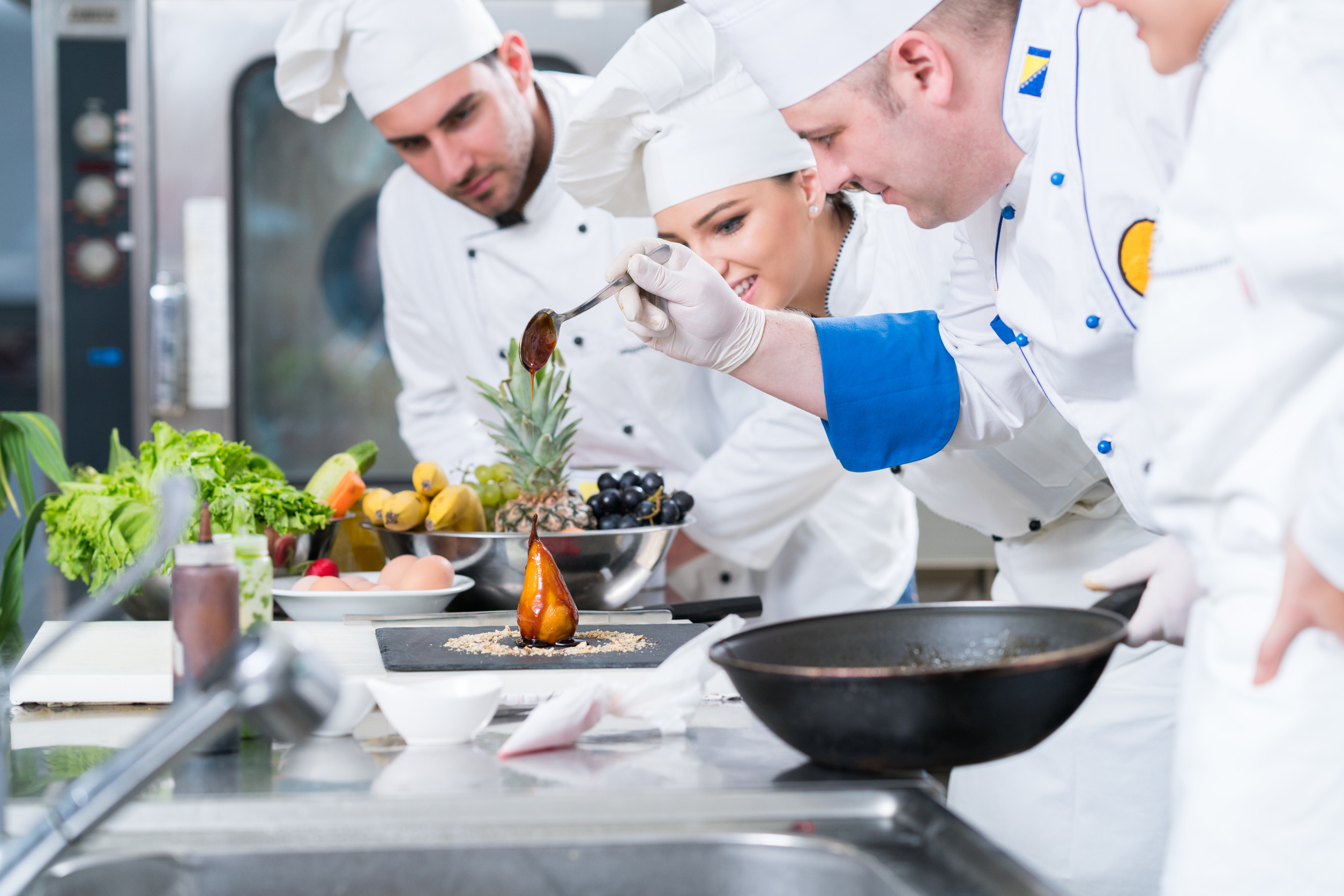 A group of professional chefs in white uniforms and hats working together in a commercial kitchen. One chef, wearing gloves and a blue-trimmed uniform, is carefully drizzling sauce over a plated caramelized pear while the others observe and smile.